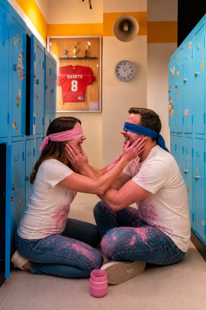 Couple réalisant un gender reveal dans un décor de vestiaires américains, les yeux bandés et couverts de peinture rose, découvrant ensemble le sexe de leur futur bébé dans une ambiance ludique et complice.