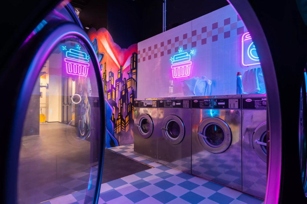 Décor de laverie automatique (vu de l'intérieur d'un sèche linge) néon avec machines à laver industrielles, éclairages colorés et ambiance urbaine rétro-futuriste idéale pour shootings photo créatifs.
