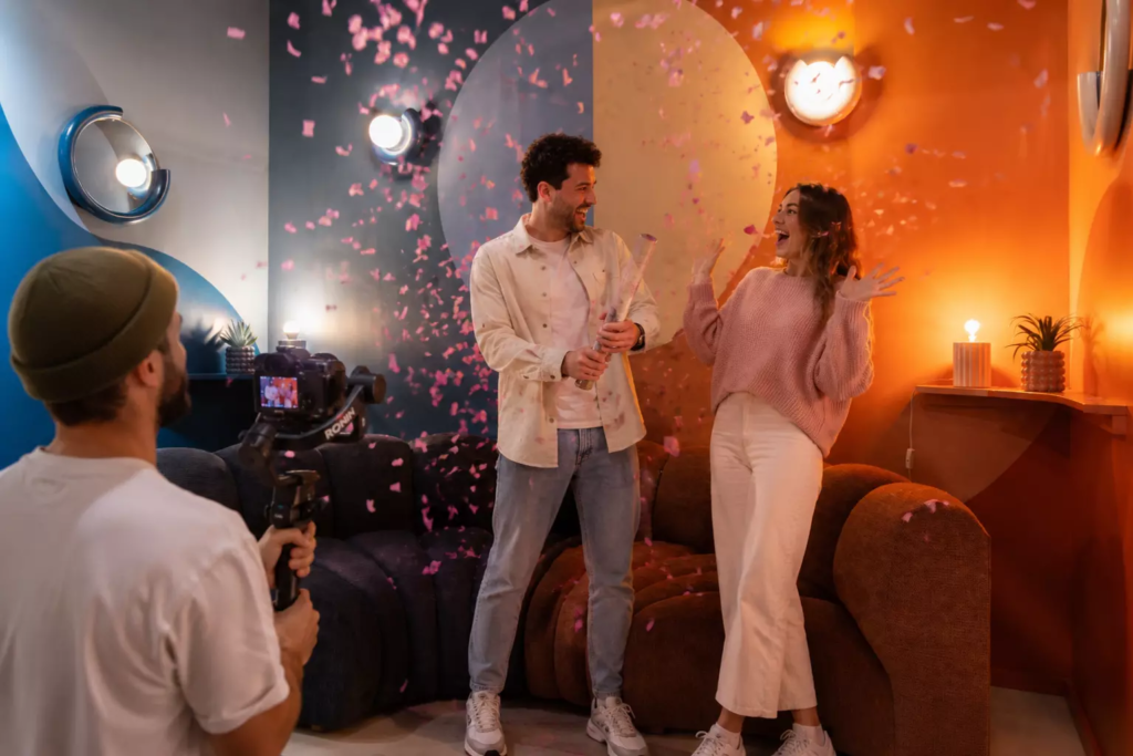Couple célébrant un gender reveal avec une pluie de confettis roses dans un décor design bicolore bleu et orange, tandis qu’un vidéaste professionnel filme leur réaction pleine de joie et de surprise.