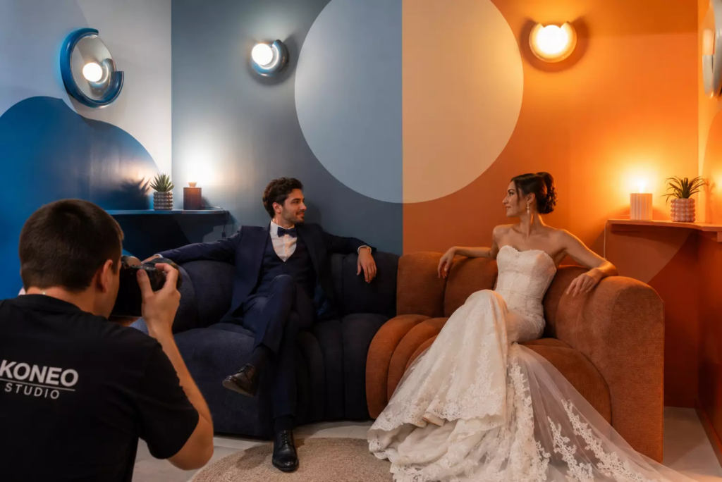 Couple de mariés réalisant un shooting photo créatif dans un décor bicolore bleu et orange, chacun installé sur une moitié de canapé assortie à son univers, tandis qu’un photographe professionnel capture leur échange de regards dans une composition graphique et contemporaine.