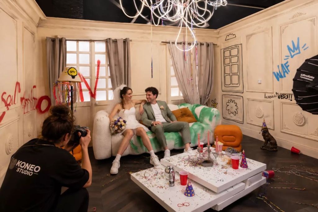 Couple de jeunes mariés réalisant un shooting photo dans un décor immersif façon appartement après soirée, assis sur un canapé design au milieu de confettis et d’accessoires festifs, tandis qu’une photographe professionnelle capture leur complicité dans une ambiance décontractée et créative.