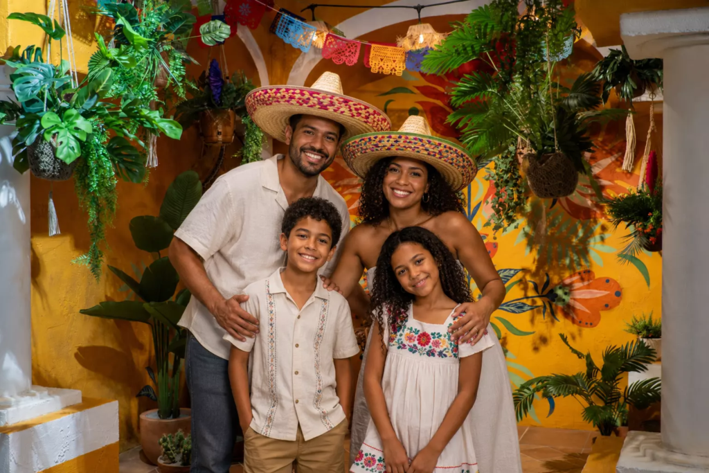 Famille de quatre personnes posant dans un décor immersif inspiré du Mexique, entourée de végétation tropicale, de couleurs chaleureuses et de décorations traditionnelles, vêtue de tenues estivales et coiffée de sombreros pour une photo souvenir pleine de bonne humeur.
