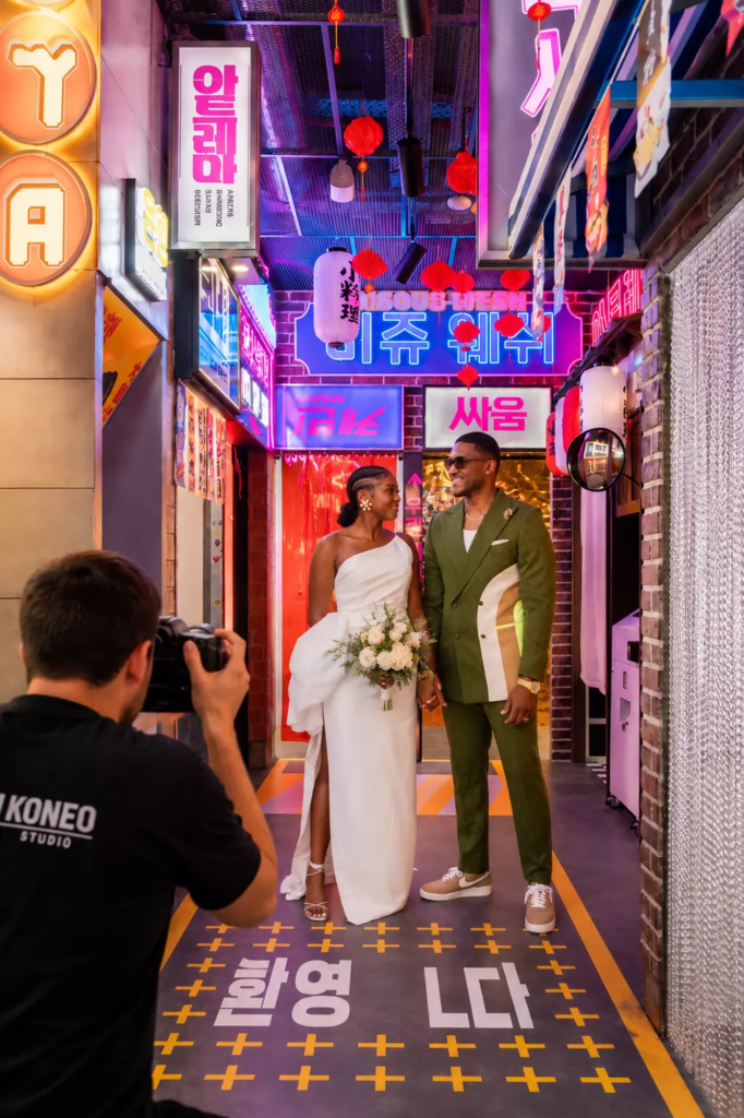 Couple de mariés réalisant un shooting photo dans une ruelle immersive inspirée de Séoul, entourée d’enseignes lumineuses et de néons colorés, tandis qu’un photographe professionnel capture leur complicité dans une ambiance urbaine contemporaine et décalée.