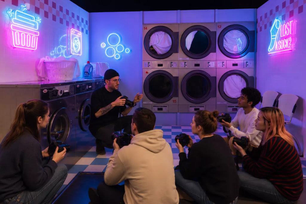 Workshop de photographie dans un décor immersif de laverie rétro éclairée aux néons, où un photographe professionnel forme un groupe d’étudiants en photo équipés de leurs appareils, dans le cadre d’un cours pratique de prise de vue.
