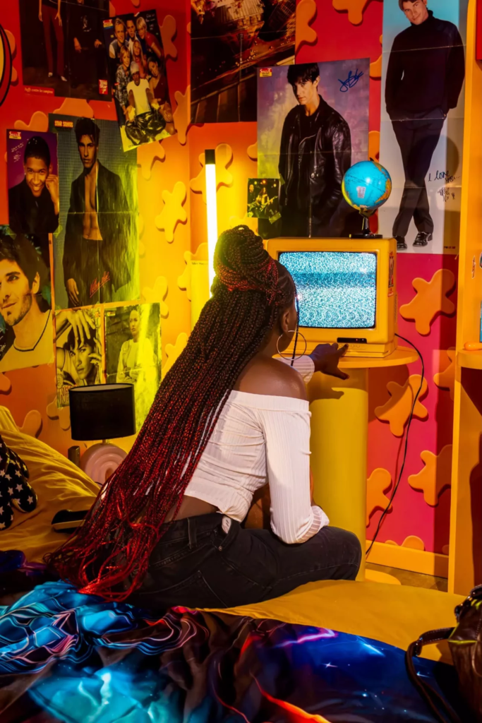 Femme assise dans une chambre immersive aux couleurs orange et jaune, face à une télévision rétro entourée de posters.