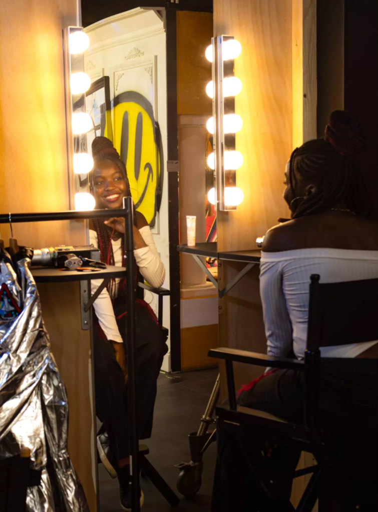 Femme souriante devant un miroir de loge éclairé par des ampoules, dans les coulisses d’un shooting photo.