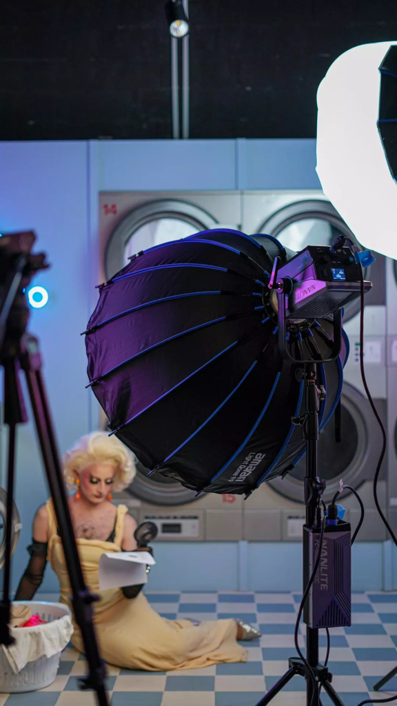 Coulisses d’un shooting photo dans une laverie rétro, avec éclairages de studio et modèle en robe jaune assise devant des machines à laver.