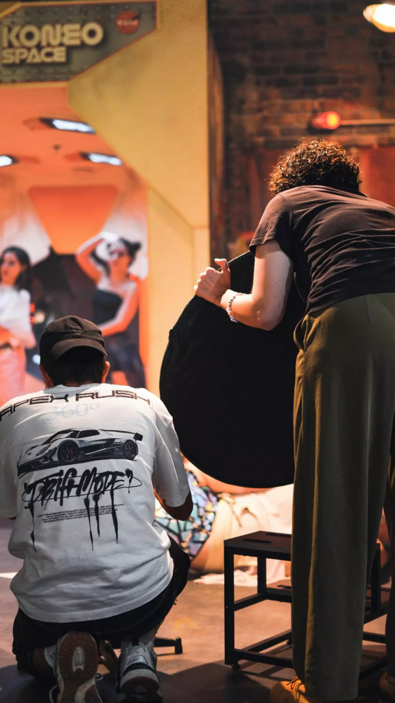 Coulisses d’un shooting photo dans un décor immersif rétrofuturiste, avec équipe technique au premier plan et modèles posant en arrière-plan.