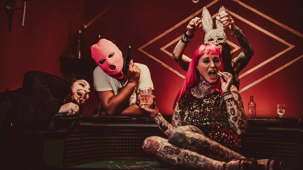 Groupe de quatre personnes posant autour d’une table de casino dans un décor rouge, avec masques, tatouages et accessoires extravagants.