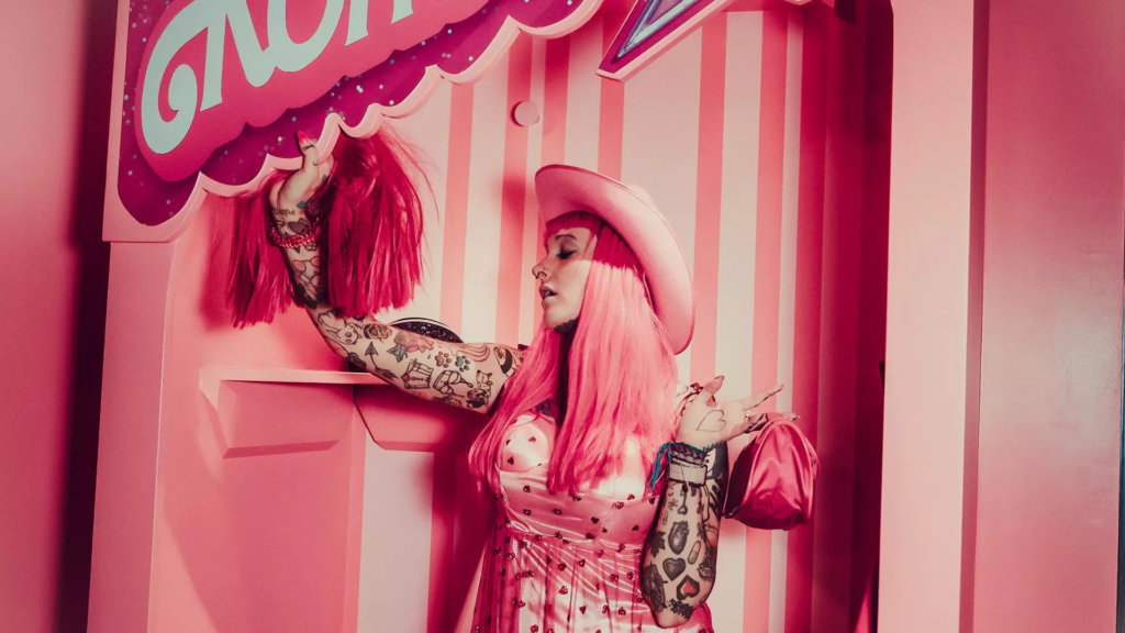Femme tatouée aux cheveux roses et chapeau de cowgirl posant dans un décor monochrome rose aux motifs rayés.