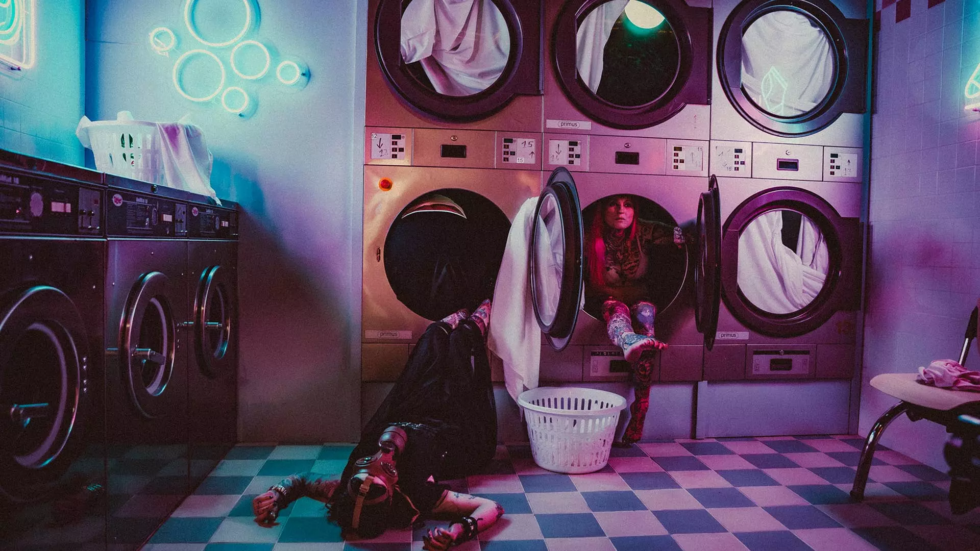 Deux personnes posant dans une laverie rétro immersive, l’une allongée au sol et l’autre assise dans un sèche-linge, sous des lumières néon roses et bleues.
