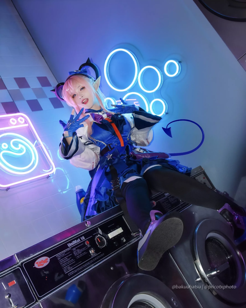Cosplayeuse aux cheveux roses en costume bleu posant sur une machine à laver dans un décor de laverie éclairé aux néons.