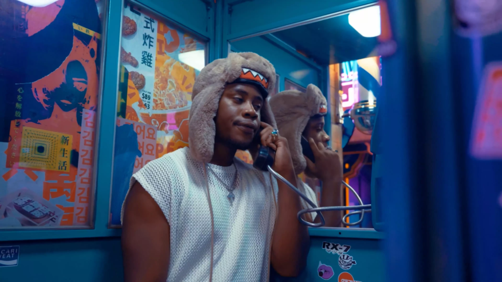 Jeune homme portant un bonnet en fausse fourrure téléphone depuis une cabine rétro aux néons, dans un décor urbain coloré d’inspiration asiatique.
