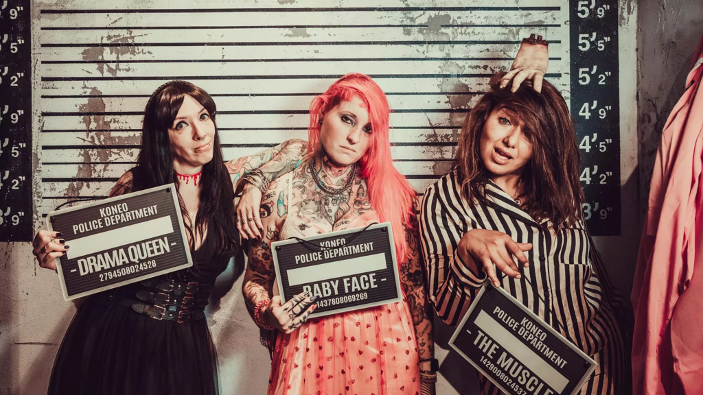 Trois femmes costumées posant devant un mur de photos d’identité judiciaire, tenant des pancartes de police dans une mise en scène décalée.