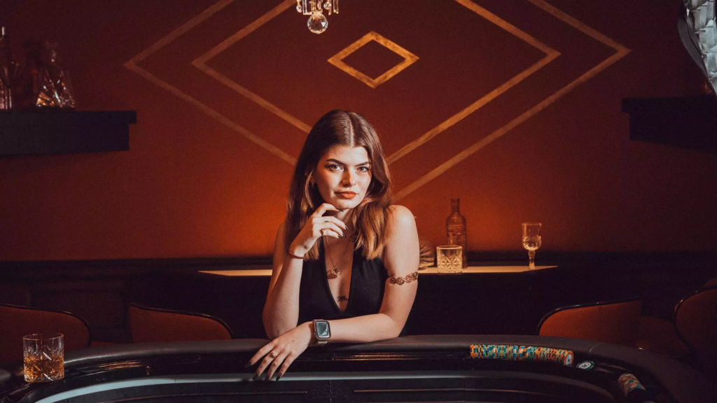 Femme posant derrière une table de casino avec jetons de poker, dans un décor élégant aux tons rouges et dorés.