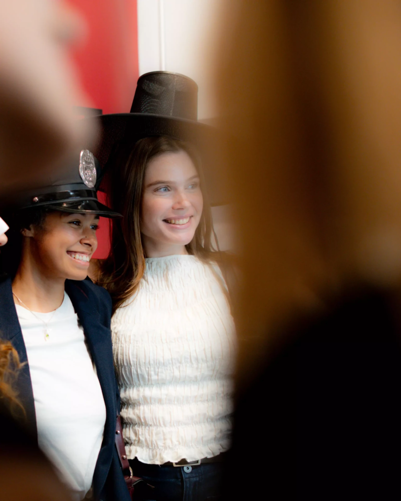 Texte alternatif : Deux jeunes femmes souriantes posent côte à côte devant un appareil photo, coiffées d’accessoires amusants, dont un chapeau haut-de-forme et une casquette de police. Photographiée à travers d’autres participants flous au premier plan, la scène restitue l’ambiance spontanée et conviviale d’une animation photo lors d’un événement.