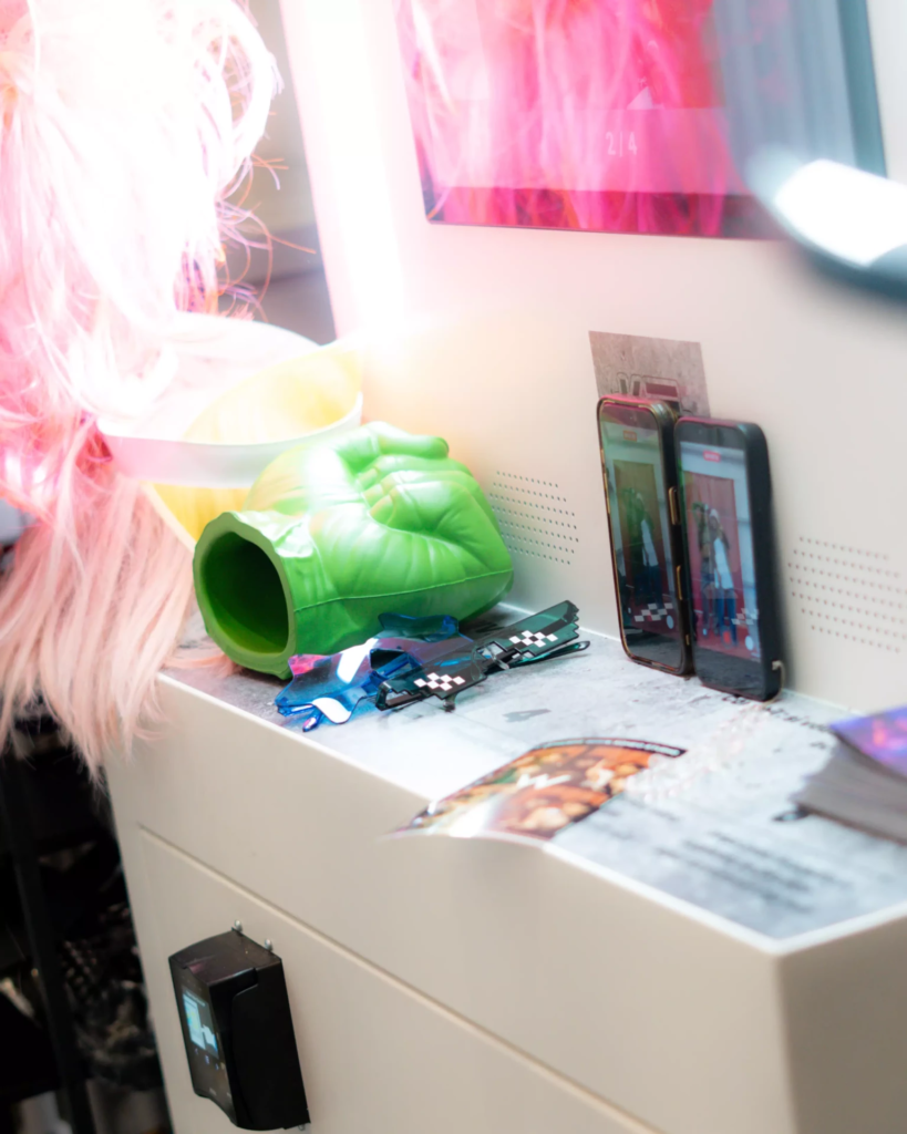 Texte alternatif : Gros plan sur un photomaton en cours d’utilisation, avec plusieurs accessoires disposés devant l’écran : perruque rose, poing vert géant et lunettes fantaisie. Deux smartphones posés contre la machine affichent la scène photographiée, illustrant une expérience photo ludique, interactive et pensée pour être facilement partagée sur les réseaux sociaux.