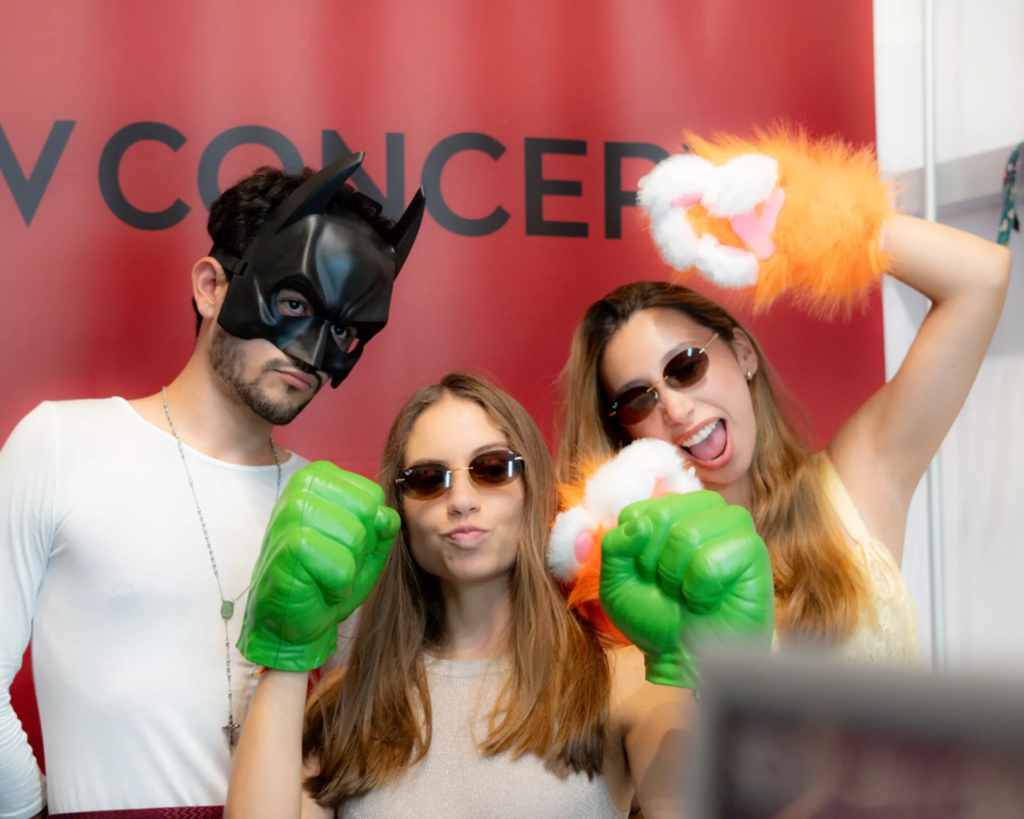 Texte alternatif : Trois amis prennent la pose devant un photomaton avec des accessoires décalés : masque noir à oreilles pointues, lunettes de soleil, poings verts géants et pattes orange en peluche. Une scène joyeuse et spontanée qui met en avant l’aspect ludique et convivial de l’expérience photo.
