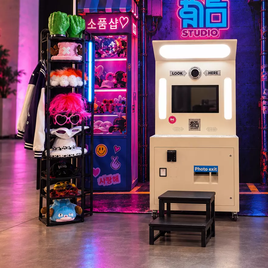 Texte alternatif : Photomaton blanc installé dans un décor immersif aux néons roses et bleus, inspiré de l’univers K-pop et des rues de Séoul. À côté, un présentoir rassemble de nombreux accessoires amusants, comme des perruques, lunettes, chapeaux et poings géants, tandis qu’un petit marchepied permet aux visiteurs de se positionner facilement devant l’appareil.