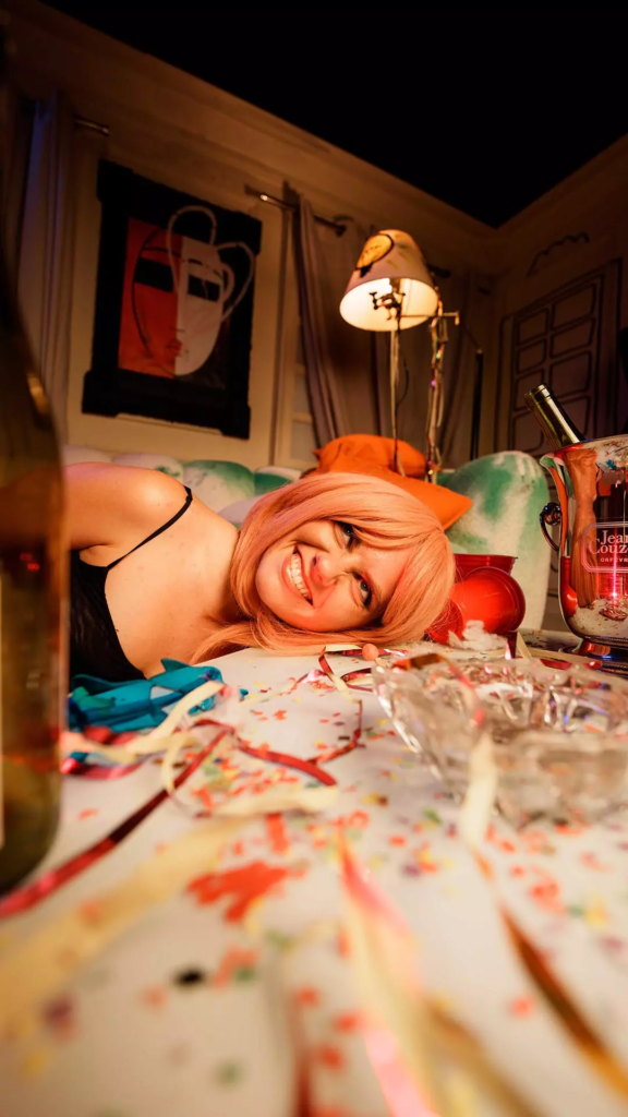 Femme aux cheveux roses souriant, la tête posée sur une table couverte de confettis et d’accessoires de fête dans un salon coloré.