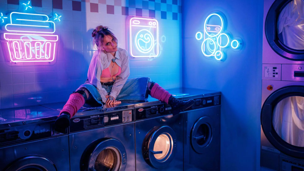 Femme posant sur des machines à laver dans une laverie rétro immersive éclairée par des néons roses et bleus.