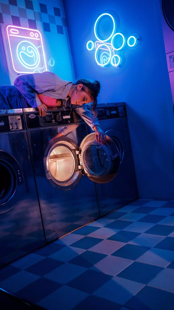 Femme penchée sur une machine à laver ouverte dans une laverie rétro immersive éclairée par des néons bleus et roses.
